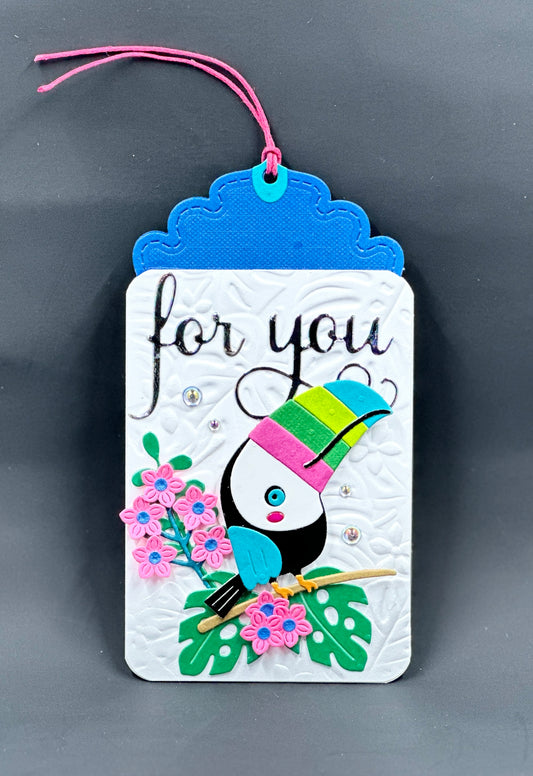 Adorable gift tag featuring a cute little Tucan sitting on a branch adorned with flowers and tropical leaves in front of an embossed background.